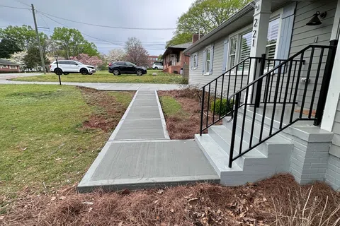 Newly poured concrete pathway leading to the steps of a gray house with black railings and manicured yard.