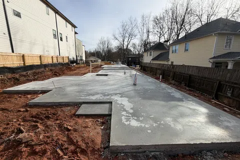 Freshly poured concrete foundation slab at a residential construction site with surrounding houses and bare trees.