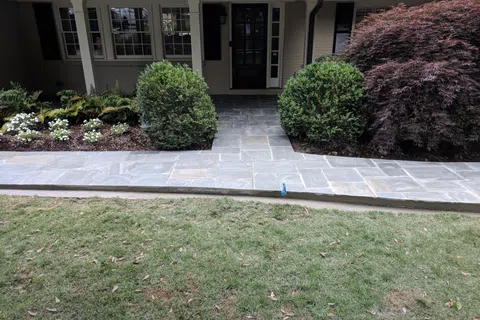 Front porch with stone pathway, green shrubs, white flowers, and a Japanese maple tree beside the house.