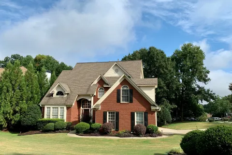 Two-story brick and beige siding house with well-maintained lawn and surrounding trees under blue sky.