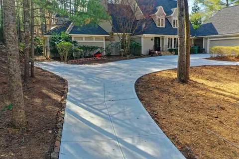 Curved concrete driveway leading to a large suburban house surrounded by trees and landscaped yard.