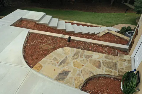 Outdoor stone patio with surrounding bare soil, concrete walkway, and stairway in a garden setting.