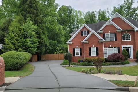 Newly paved gray concrete driveway leading to a red brick two-story house with black shutters and surrounding greenery.