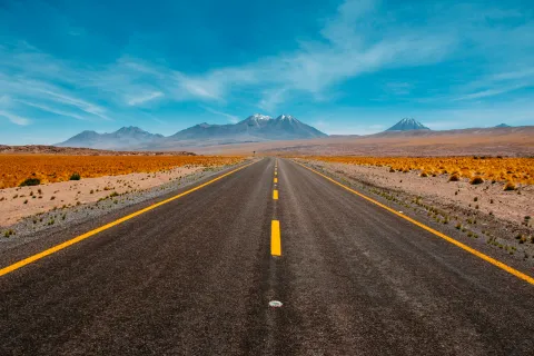 Straight pavement road with yellow lines leading to distant mountains under blue sky in arid landscape.