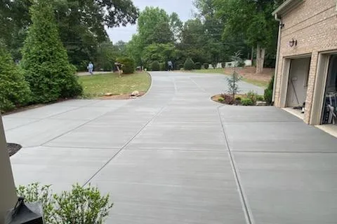 Wide concrete driveway next to brick house with greenery and trees in the background on a cloudy day