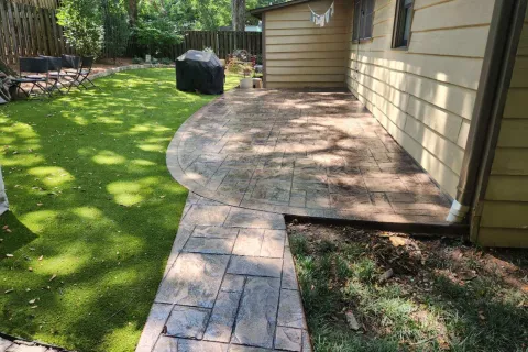 A backyard patio with stamped concrete and artificial grass, surrounded by trees and a grill.