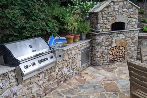 Outdoor kitchen with stainless steel grill, stone pizza oven, firewood storage, and potted plants on stone countertop.