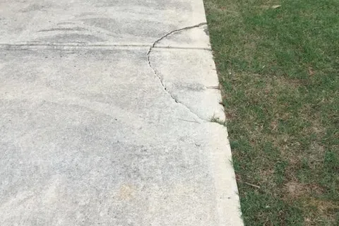 Cracked concrete driveway with basketball hoop on a grassy area, showing signs of wear and tear.