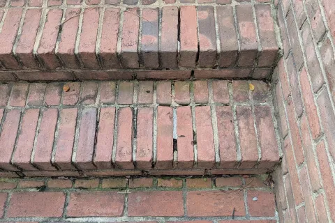 Close-up of weathered red brick steps with cracks and worn mortar, bordered by brick walls on one side.