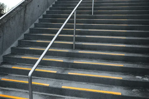 Concrete outdoor stairs with yellow safety strips and a central metal handrail under a cloudy sky.