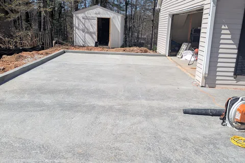 Freshly poured concrete driveway next to a white garage and a small white storage shed surrounded by trees.