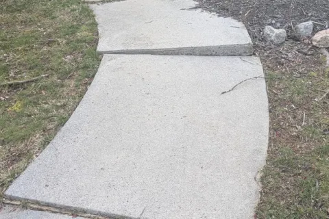 Cracked concrete sidewalk winding through green grass and landscaping near stairs.
