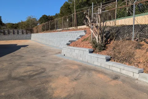 Concrete retaining walls with steps lining a dirt slope beside an asphalt area under clear blue sky.