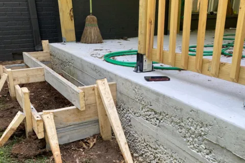 Wooden framing and freshly poured concrete foundation for outdoor stairs next to a porch with broom and hose.