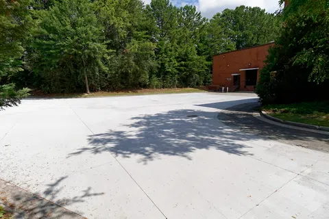 A bright, empty parking lot surrounded by lush green trees under a clear blue sky.