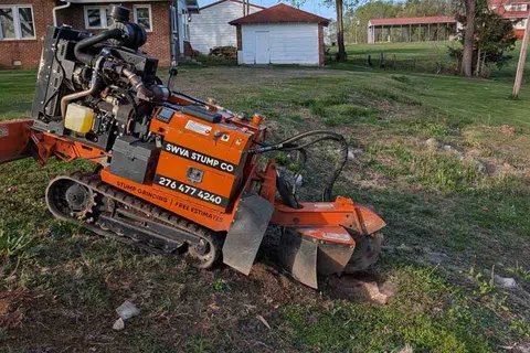 Orange stump grinding machine labeled SWVA Stump Co working on clearing a tree stump in a yard near houses.