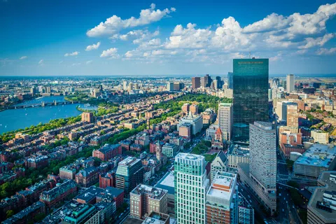 Aerial view of Boston skyline with river, parks, and skyscrapers under a partly cloudy blue sky.