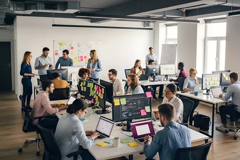 Modern open office with diverse team working at desks with computers and sticky notes on monitors and walls.