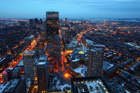 Aerial view of a city skyline at dusk with illuminated streets, tall buildings, and distant water body.