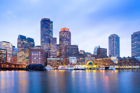 Boston waterfront skyline at dusk with illuminated skyscrapers reflecting on calm water under a clear sky.