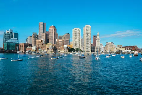 Sailboats docked in blue harbor waters with Boston city skyline and clear sky in the background.