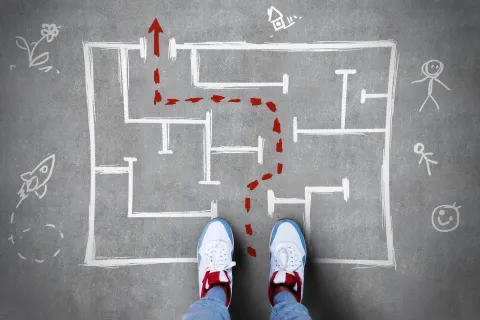 Person wearing sneakers stands before a chalk-drawn maze with a red path leading to the exit on concrete.