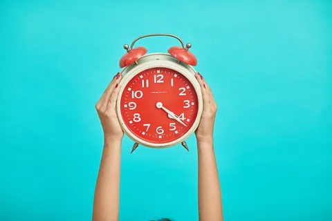 Hands holding a large red alarm clock against a bright turquoise background showing 4:20 time.