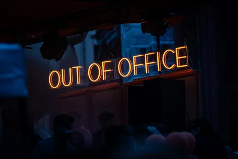 Glowing orange neon sign reading OUT OF OFFICE hanging indoors with people in dark foreground