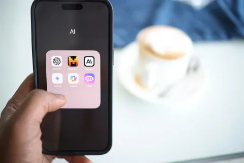 Hand holding smartphone displaying an AI app folder with ChatGPT, Mistral AI, Claude, Gemini, Copilot, and Poe icons.