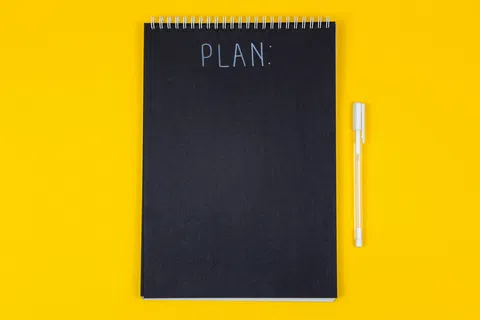 Black spiral notebook with PLAN written on cover and a white pen on a yellow background.