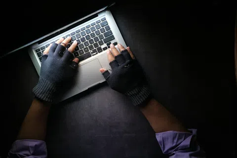 Hands wearing fingerless gloves typing on a laptop keyboard against a dark background.