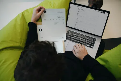 Person reviewing documents and entering information on a laptop while sitting on a green bean bag chair.