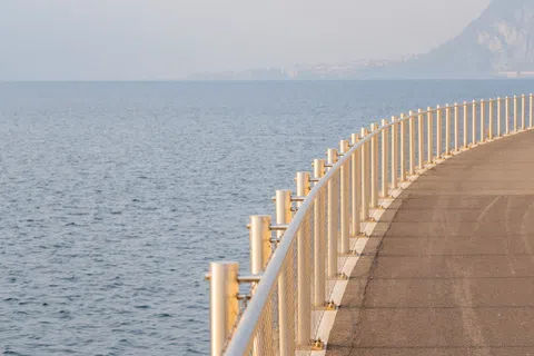 Curved waterfront pathway with metal railing beside calm sea under hazy sky and distant mountains.