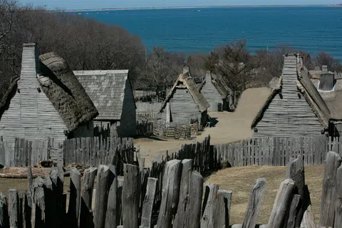 Historic village with wooden houses and thatched roofs surrounded by palisade fences near a large body of water.