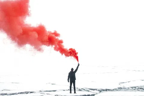 Person in black stands on icy terrain holding a red smoke flare against a bright white sky.
