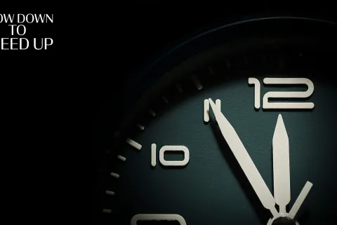 Close-up of a clock face showing the time 12:55 with the text Slow Down to Speed Up in white.