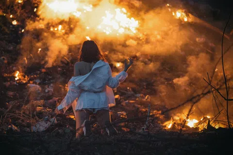 Woman in white shirt stands facing a large fire burning in a dark, outdoor environment at night.