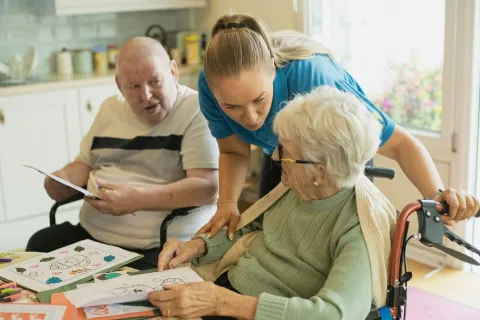 Caregiver assisting elderly woman in wheelchair with coloring activity while an elderly man observes at table.