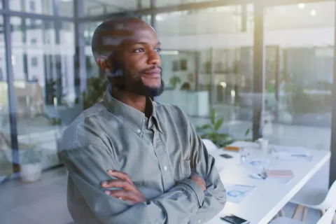 Confident young man with arms crossed looking out office window in modern workspace with natural light.