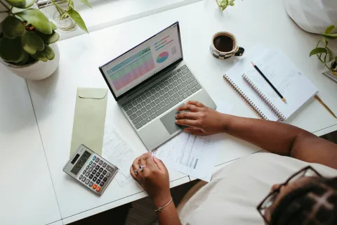 Person using laptop and calculator to review financial documents on a white desk with plants and coffee.
