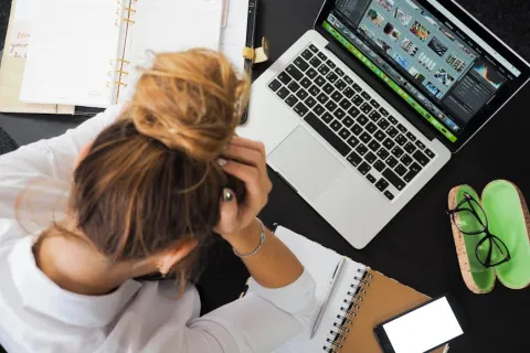 Person overwhelmed at desk with laptop, notebooks, smartphone, and eyeglasses during work or study session.