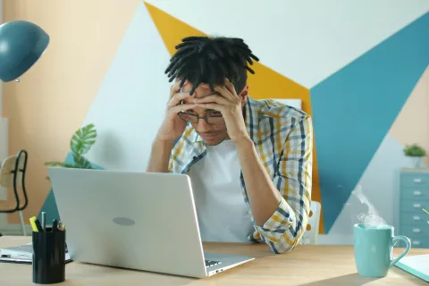 Stressed young man holding his head while working on a laptop in a colorful modern office space.
