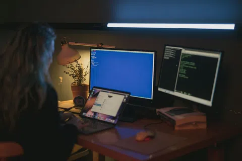 Person working on laptop and dual monitors displaying code in dimly lit workspace with desk lamp and plant.