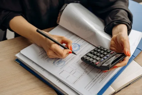 Person calculating and filling out financial documents with a calculator and pen on a wooden desk.