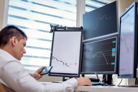 Man analyzing stock market charts on multiple computer monitors in a bright modern office setup.