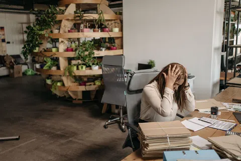 Stressed woman sitting at desk holding head with paperwork and files in a modern office with plants.
