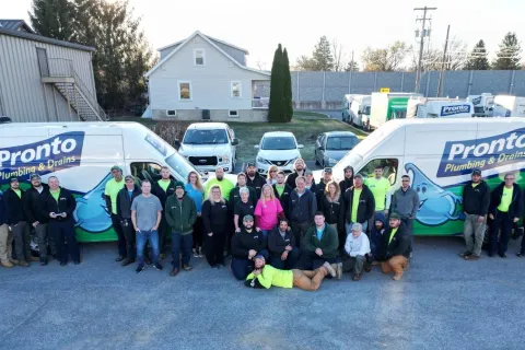 Large group of Pronto Plumbing & Drains employees posing in front of company vans in a parking lot