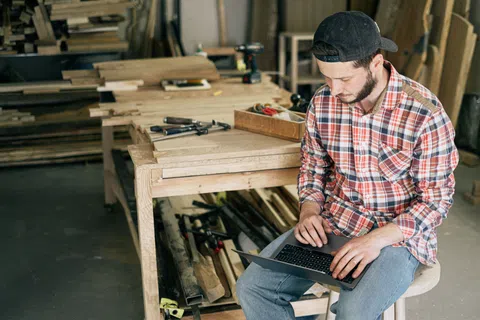 Carpenter in plaid shirt working on laptop in a woodworking shop surrounded by tools and wood materials