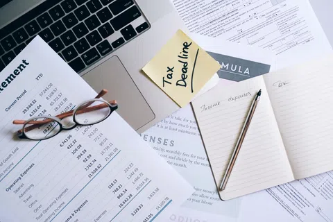 Workspace with tax documents, glasses, laptop, sticky note reading Tax Deadline, and notebook with pen