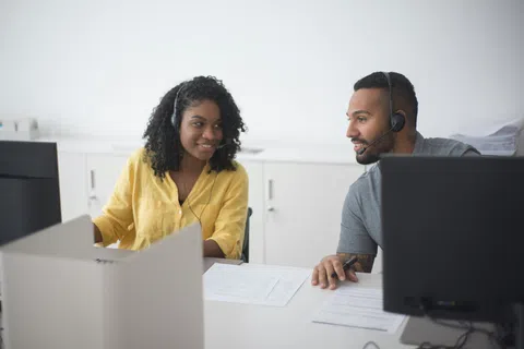 Two customer service representatives wearing headsets assisting clients at computers in an office setting.